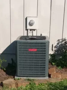 Goodman AC unit installed outside a home, placed against a light-colored wall with an electric meter mounted above it.