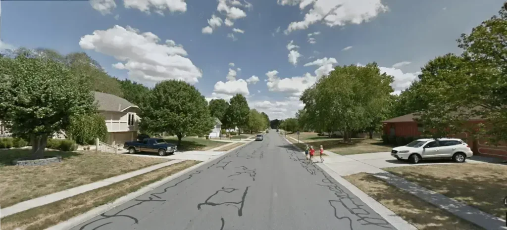 Residential street in Lee's Summit MO with suburban homes, parked cars, green trees, and kids walking on a sunny, cloudy day.