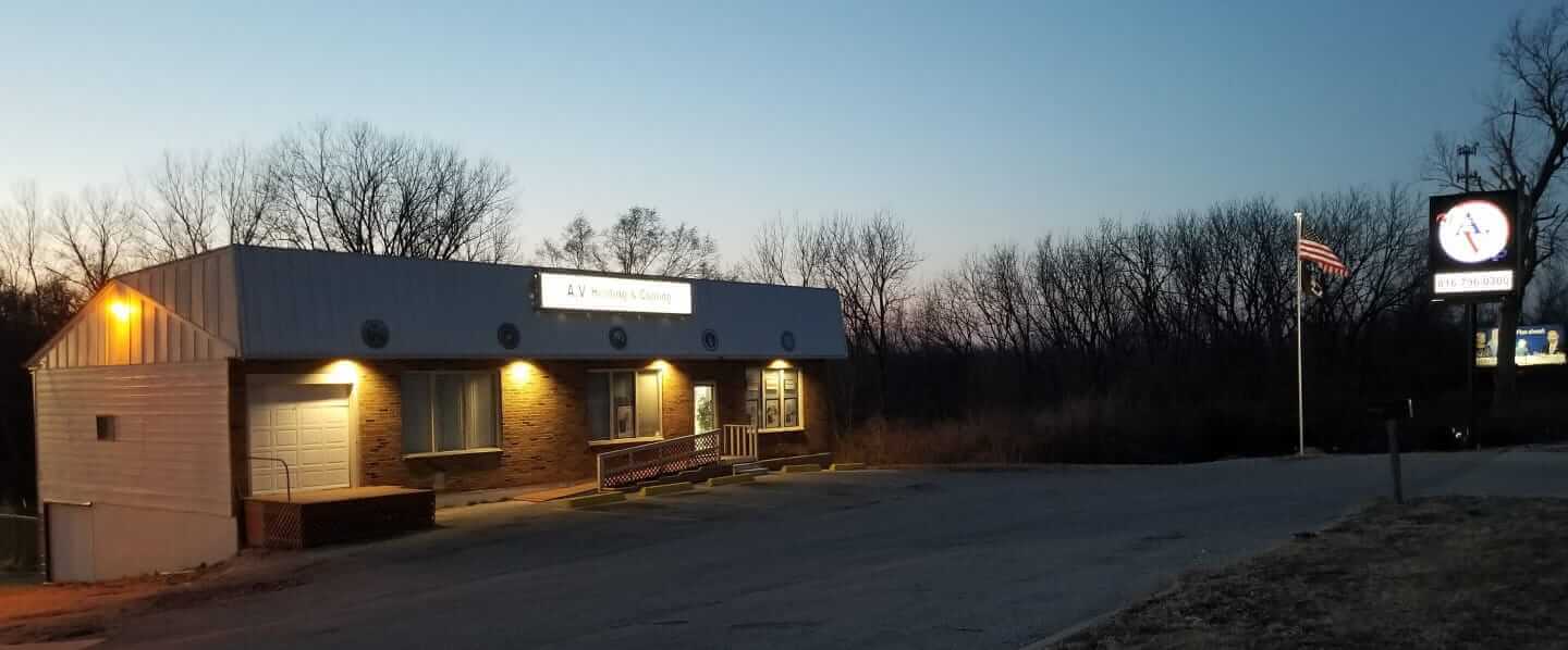 A.V. Heating & Cooling office building at dusk, with exterior lights on, an American flag, and a lit business sign by the roadside.
